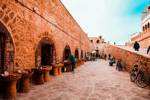 Morocco Tour Packages ,Small camel caravan trekking across orange Erg Chebbi sand dunes at sunset,.Vibrant display of colorful spices, pottery, and textiles in a bustling Marrakech souk alleyway,Historic blue fishing boats anchored on Essaouira beach with fortified city walls in background,Group of travelers enjoying traditional Moroccan mint tea and pastries inside a riad courtyard.,Luxury desert glamping tents under starry night sky in Merzouga Sahara, Morocco.